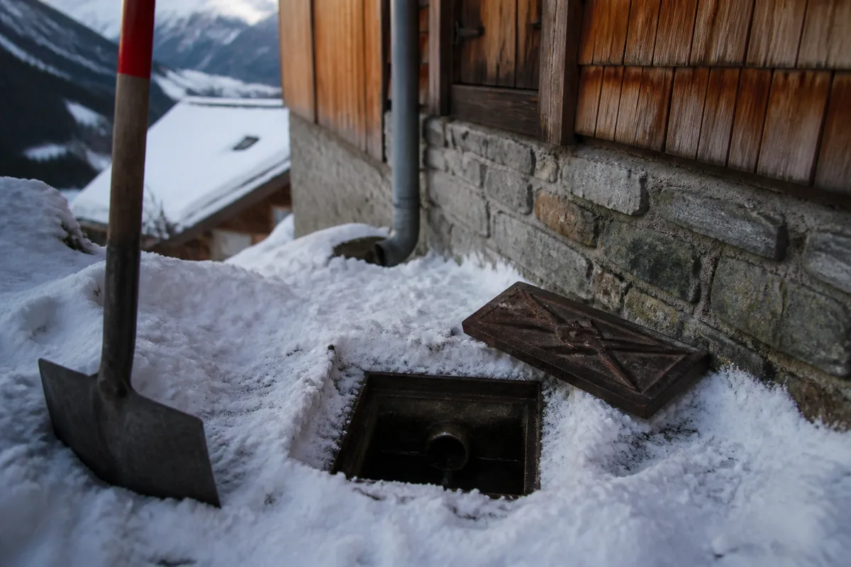 Regard de visite déneigé au pied d’un chalet, tampon de fonte posé sur la neige tassée