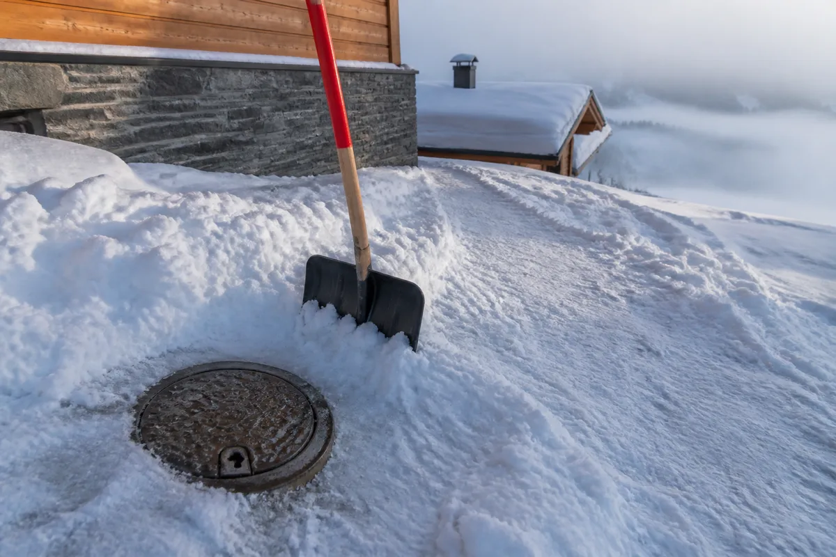Regard de compteur d’eau dégagé dans la neige tassée devant un soubassement de chalet en pierre grise